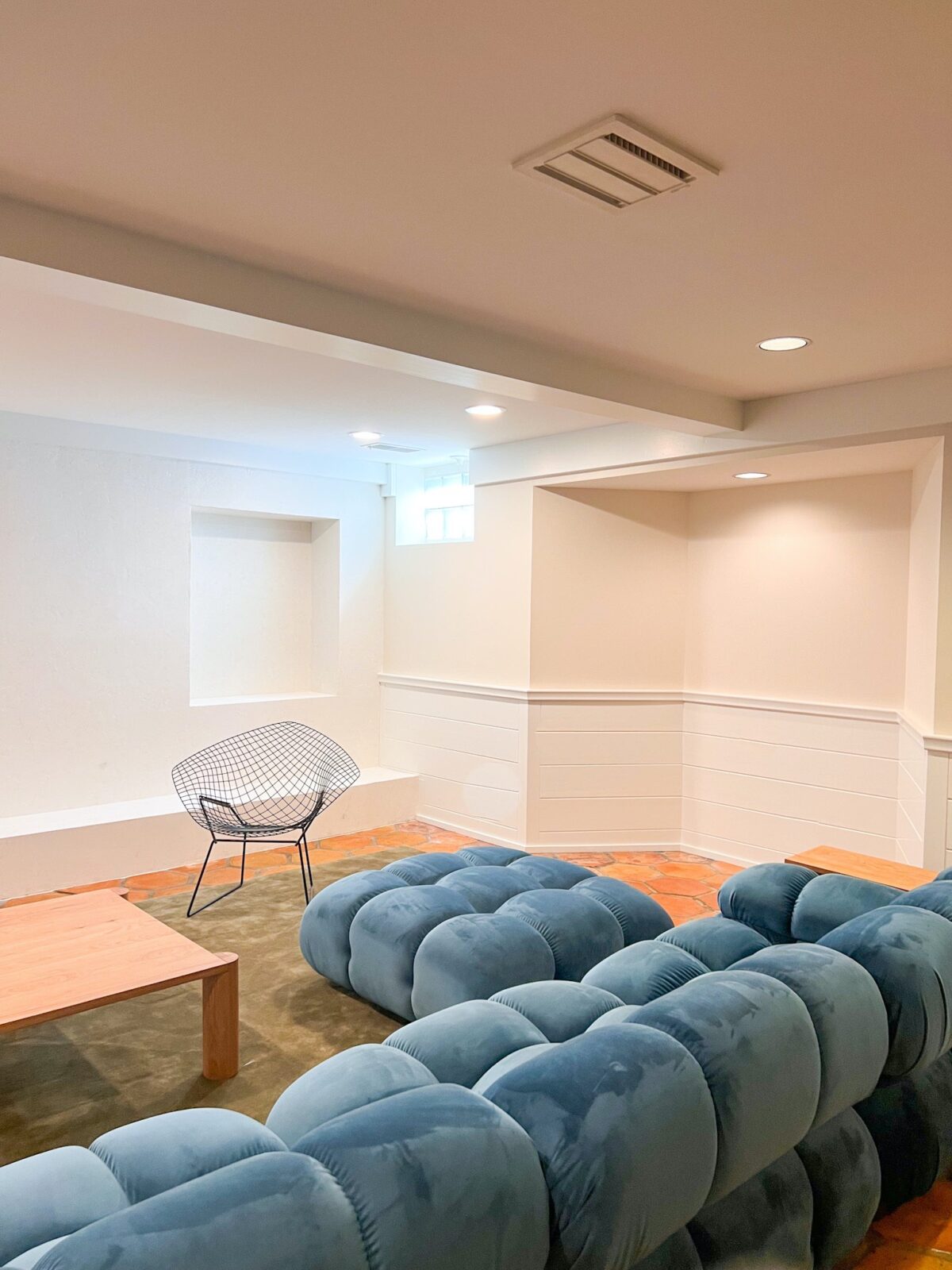 A basement family room with white and cream walls, plush green rugs, a blue modular sofa, a white oak coffee table, and vintage Bertoia diamond chairs