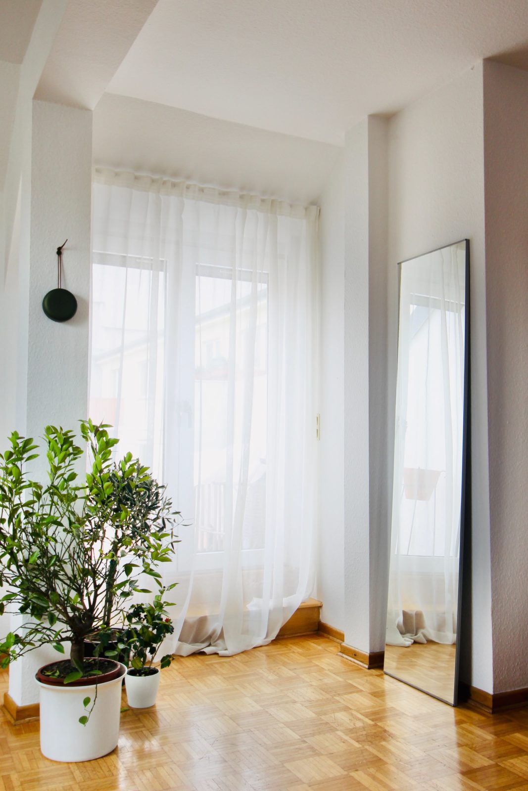 A white apartment features light brown wood floors, a floor-length mirror, plants, and breezy curtains hanging over French doors.