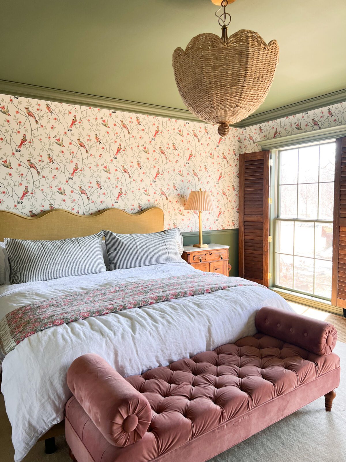 View of a bedroom, with a chaise lounge at the front of the bed. A window is letting in sunshine.