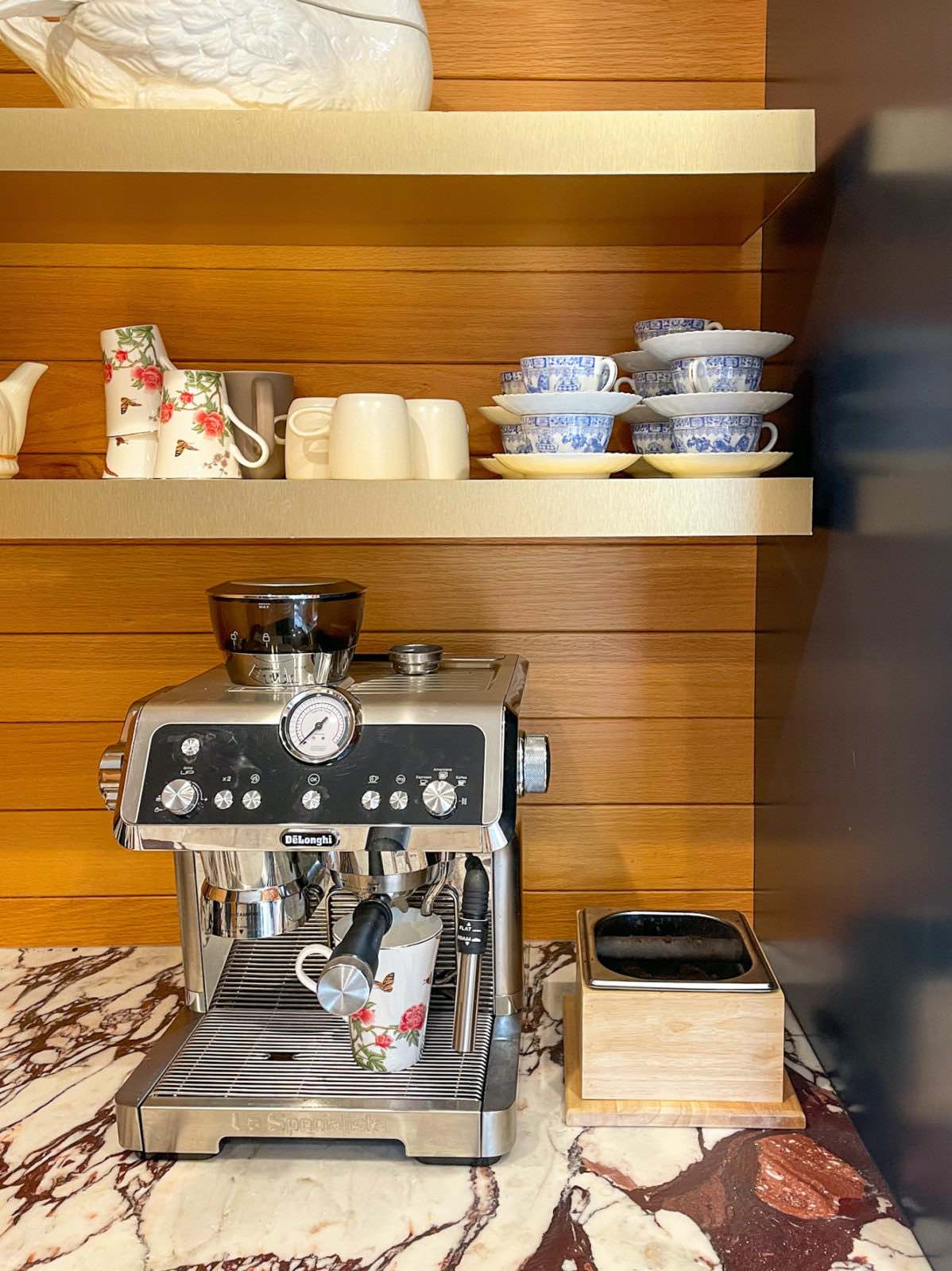 De'Longhi La Specialista Espresso Machine on a marble countertop. A wood wall is behind and 2 brass shelves are above, with coffee cups atop them.