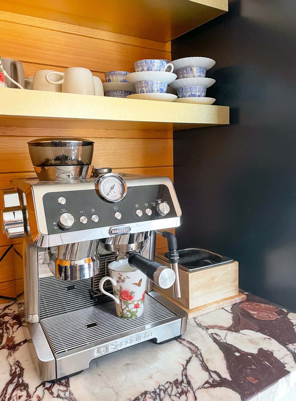 De'Longhi La Specialista Espresso Machine on a marble countertop. A wood wall is behind and 2 brass shelves are above, with coffee cups atop them.