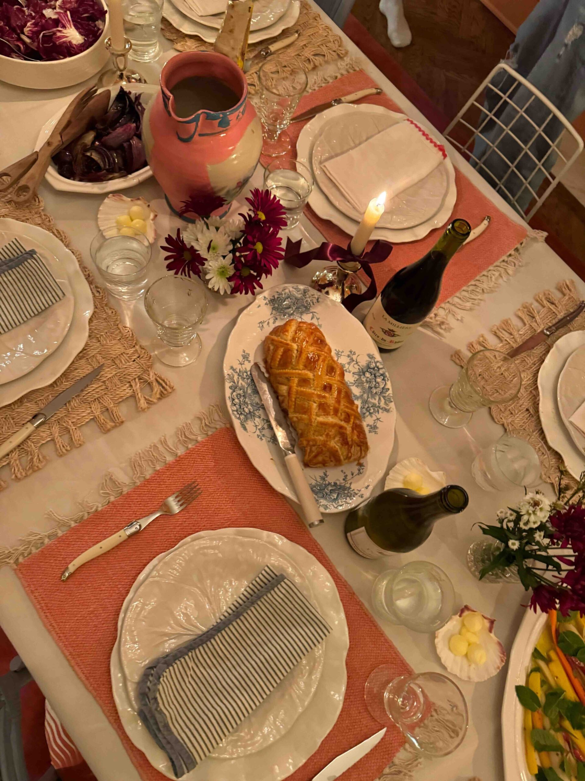 In the warm glow of candlelight, a table is set for dinner with mismatched placemats, white plates and chargers, striped napkins, and open bottles of wine