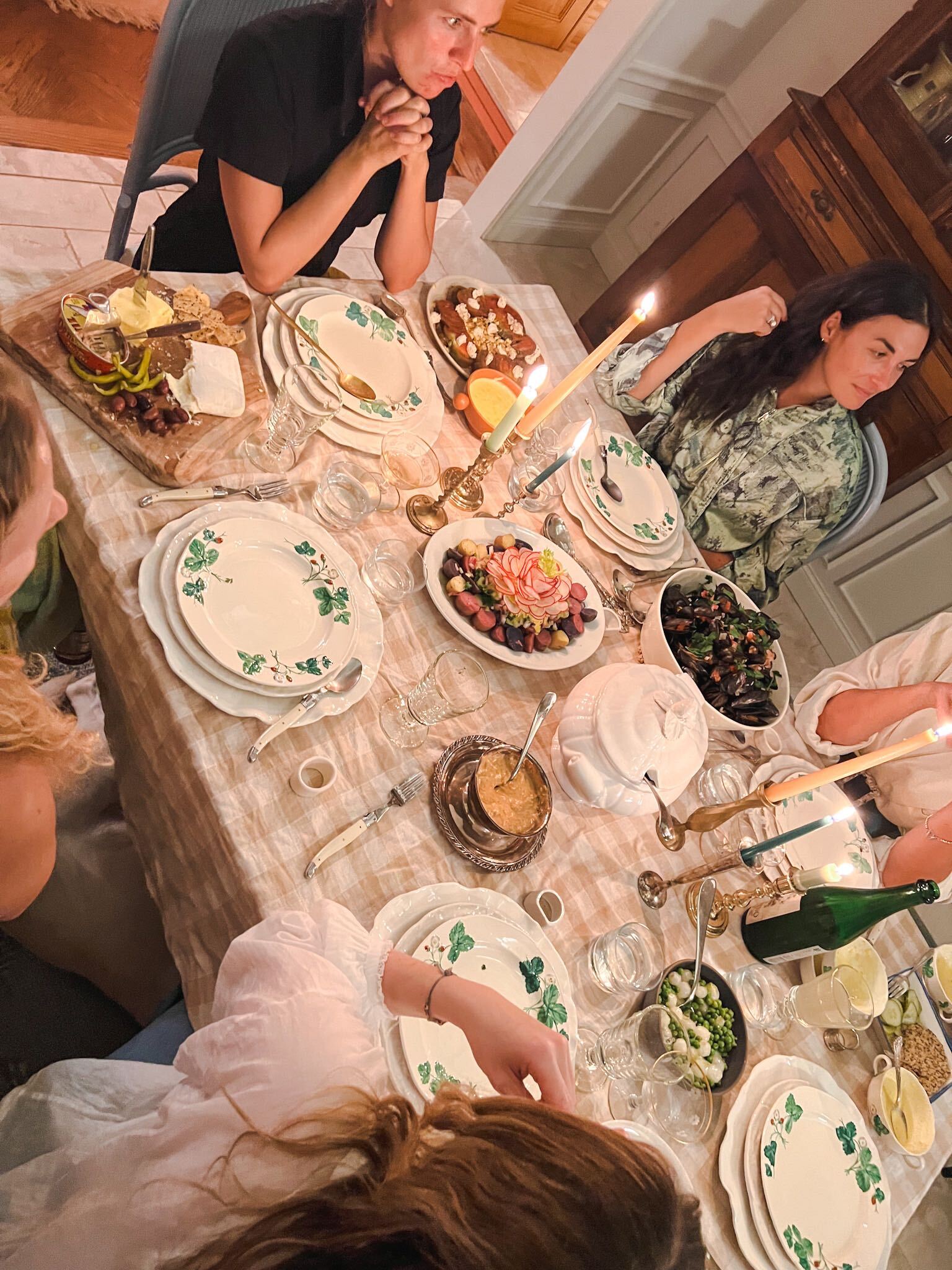 Five friends are seated around a dining table at a dinner party. The table is covered by a checkered tablecloth, place settings, lit candles, drinks, and food.