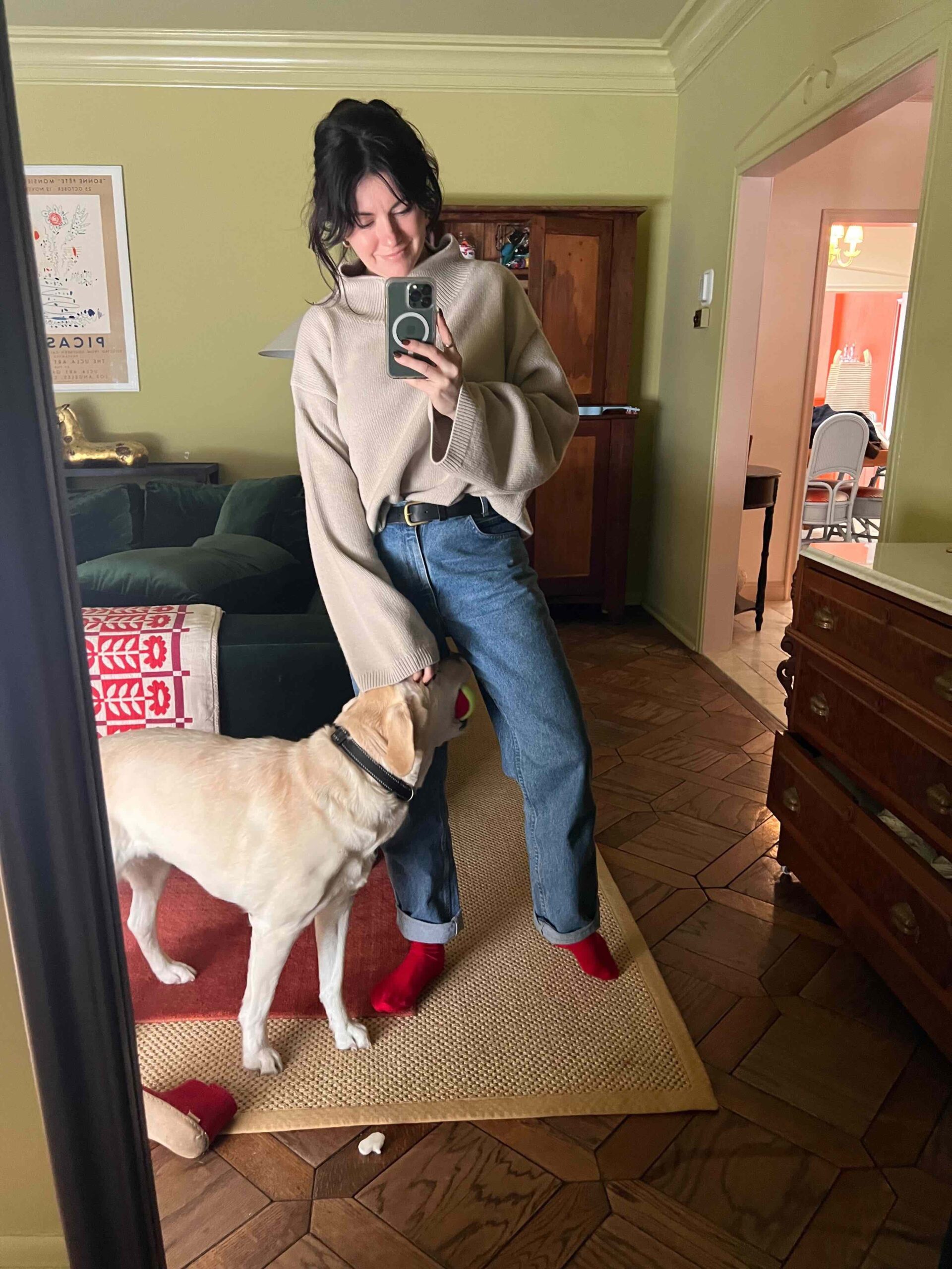 A woman wears a cozy turtleneck sweater, jeans, and red socks, while playing with her dog at home