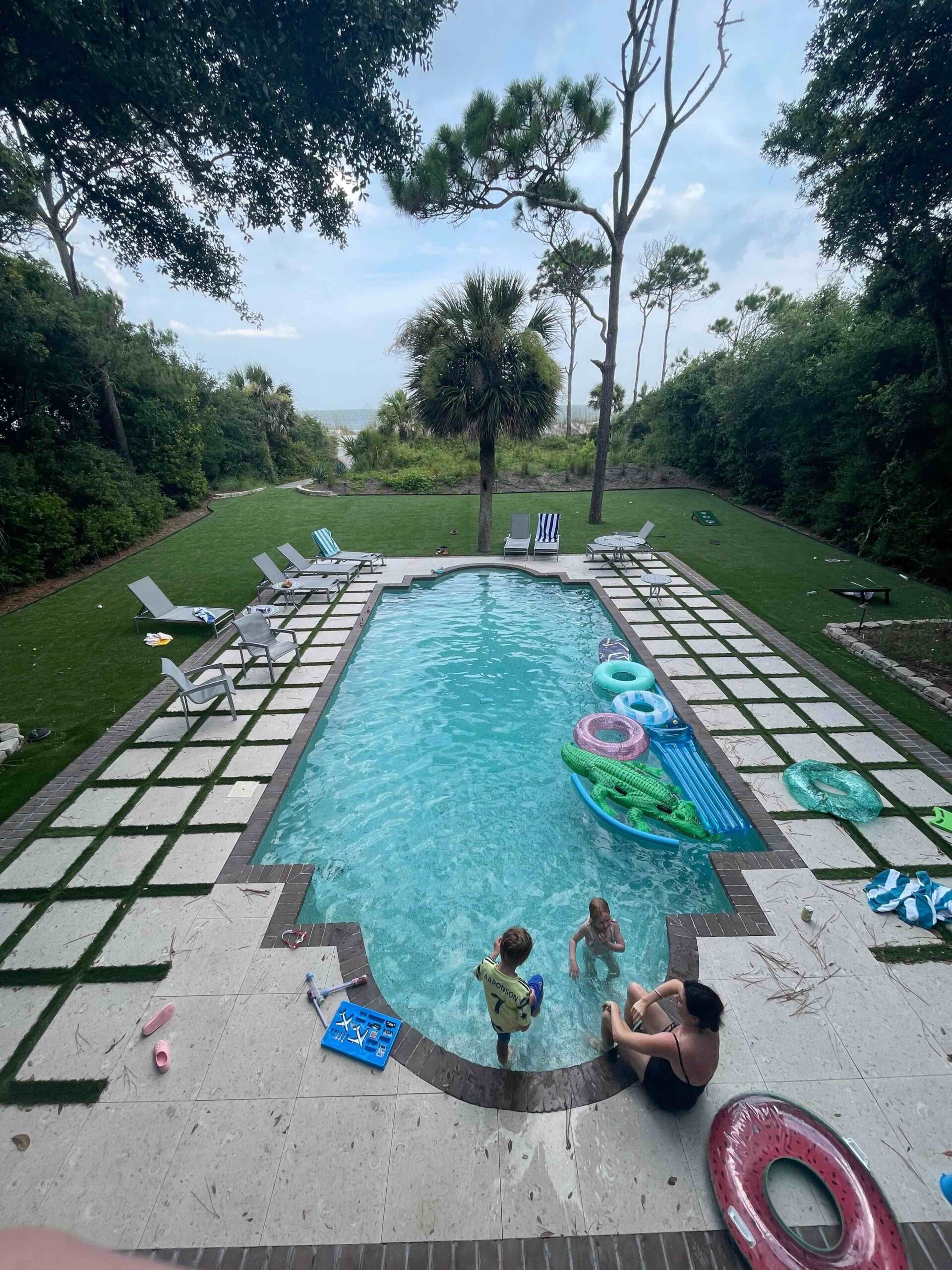 A woman and two kids are enjoying a vast backyard pool surrounded by a green lawn, with the ocean visible in the distance