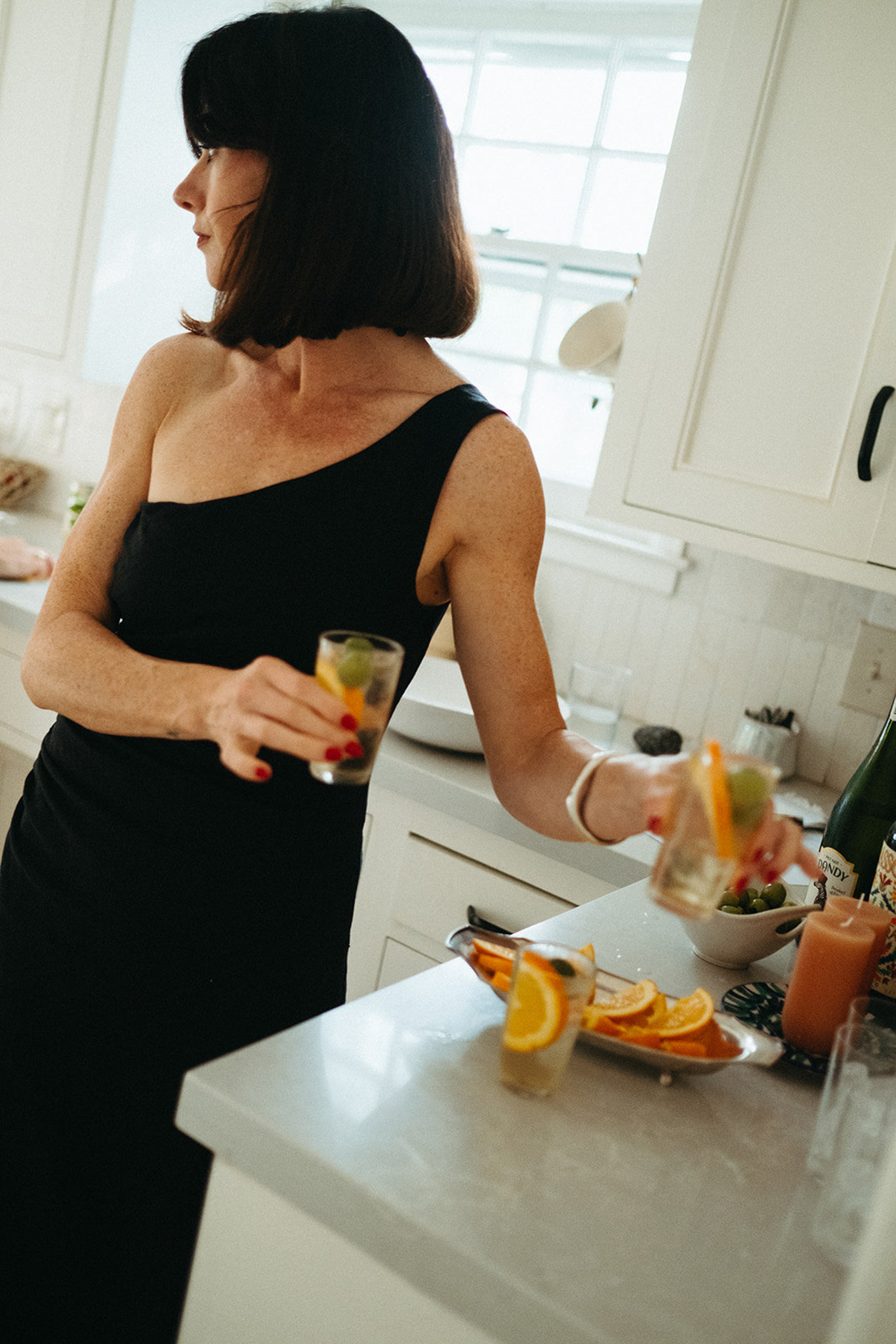 A woman wears a black one-shoulder dress. She's standing in a kitchen and holding two small vermouth spritz cocktails.