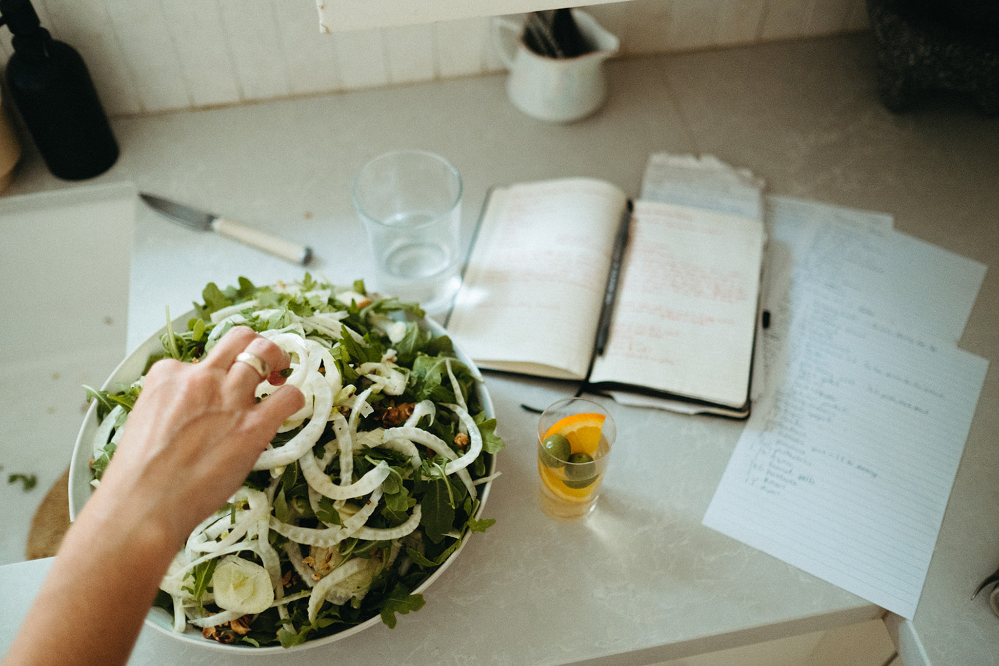 A large serving bowl filled with fresh arugula and fennel sits on a white countertop. Next to the bowl are scattered pieces of paper with recipes and ingredients lists.