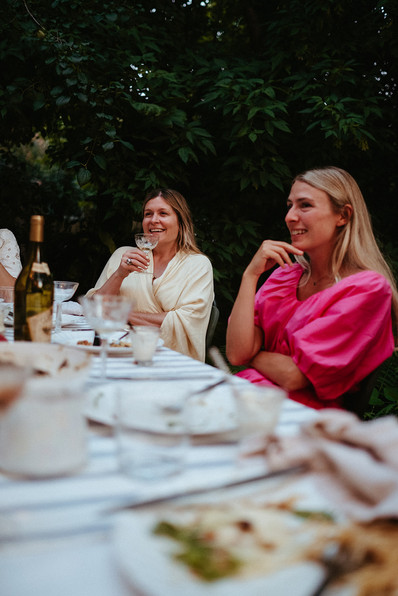 Two friends are shown sitting at an outdoor dining table at dusk, laughing, chatting, and drinking wine.