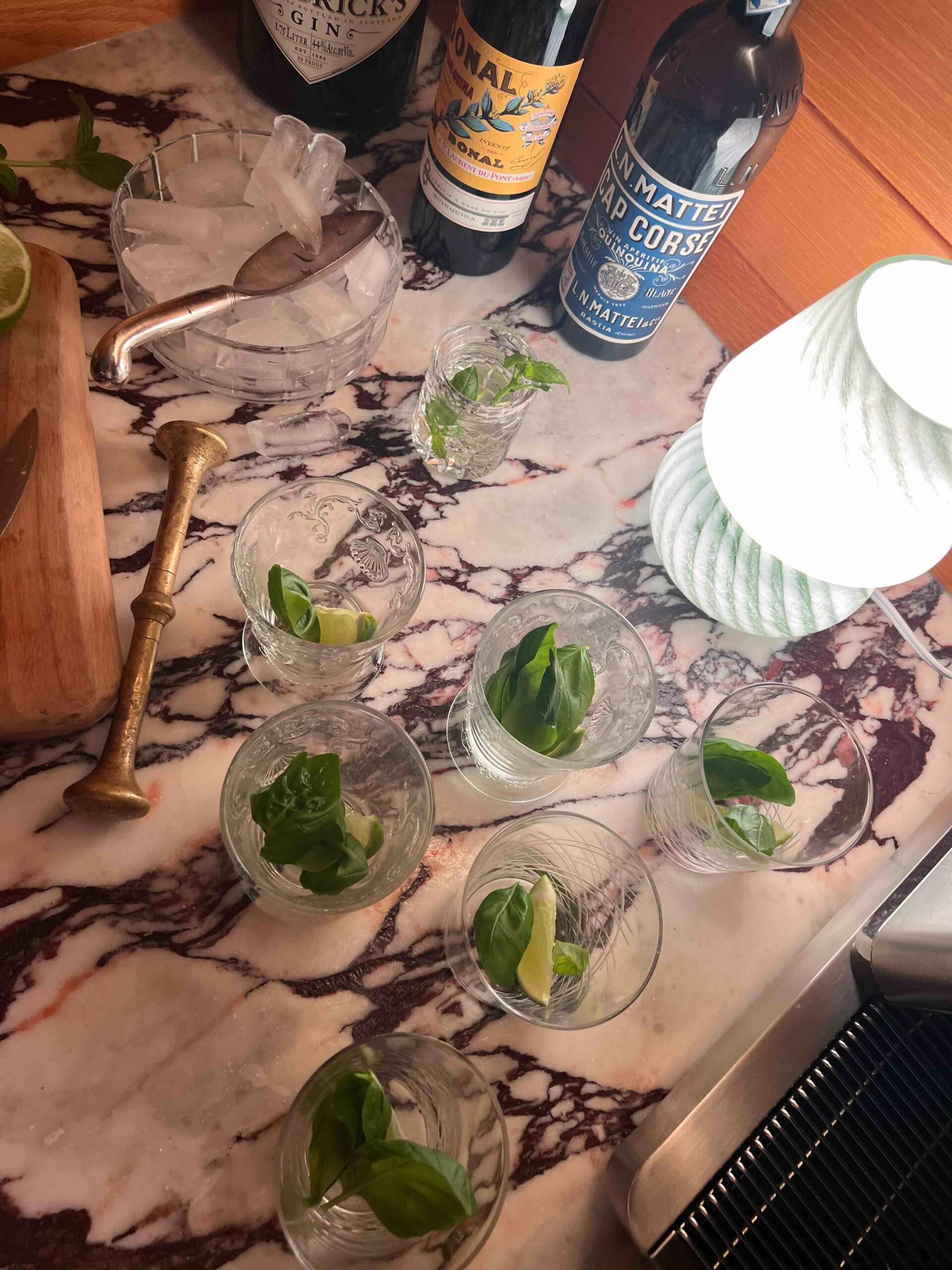 Cocktail glasses filled with mint and lime wedges are sitting on a marble countertop, with a bucket of ice and bottles of gin and liquor nearby