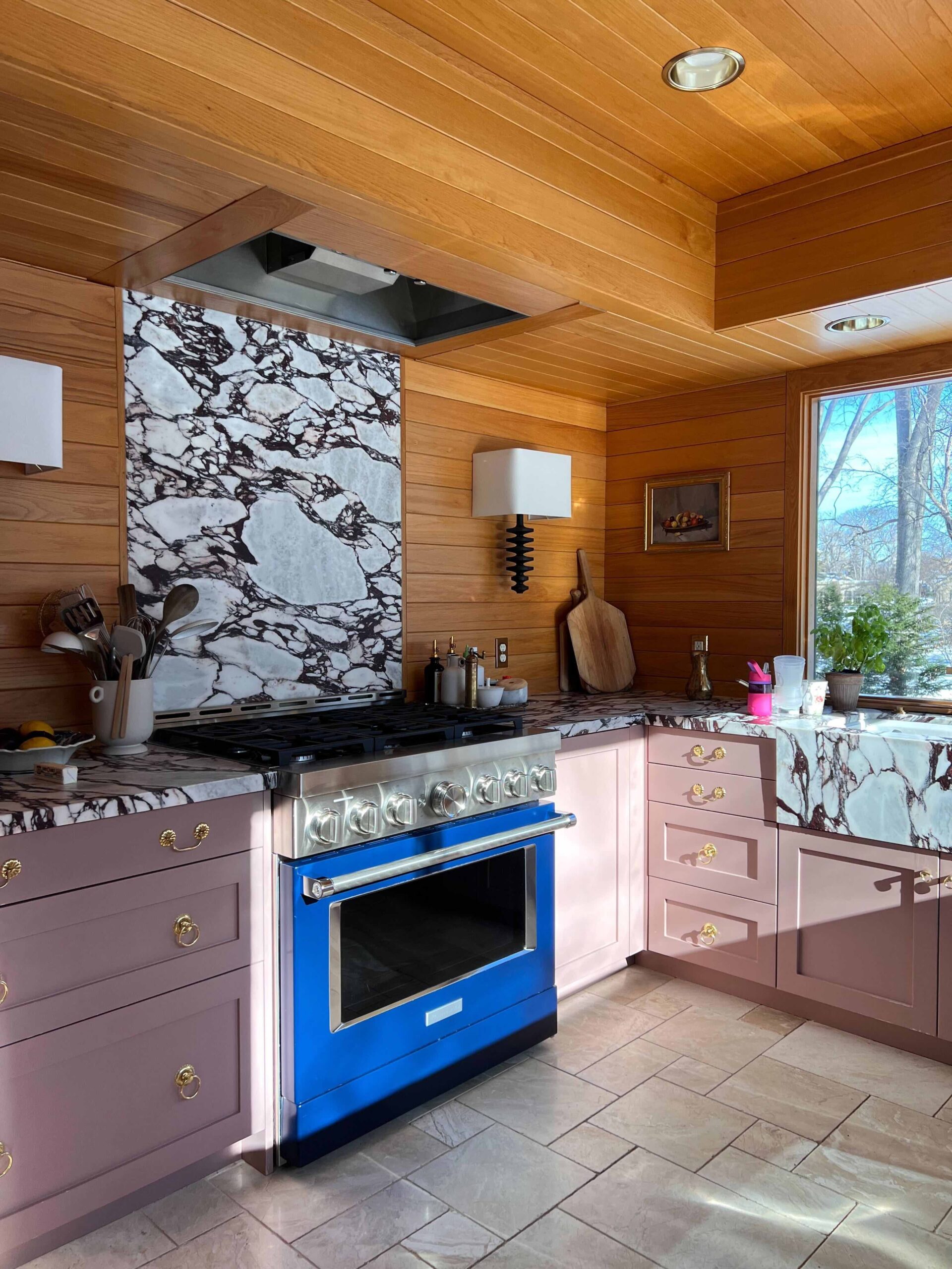 A kitchen with marble stone floors, dusty pink cabinets, marble countertops and backsplash, and a blue range gleams in the morning sunlight