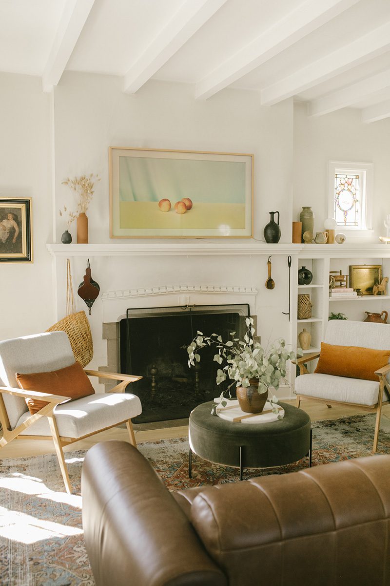 A living room has walls painted in White Dove by Benjamin Moore. A fireplace anchors the cozy space, which is decorated in neutral hues with pops of deep orange and green.