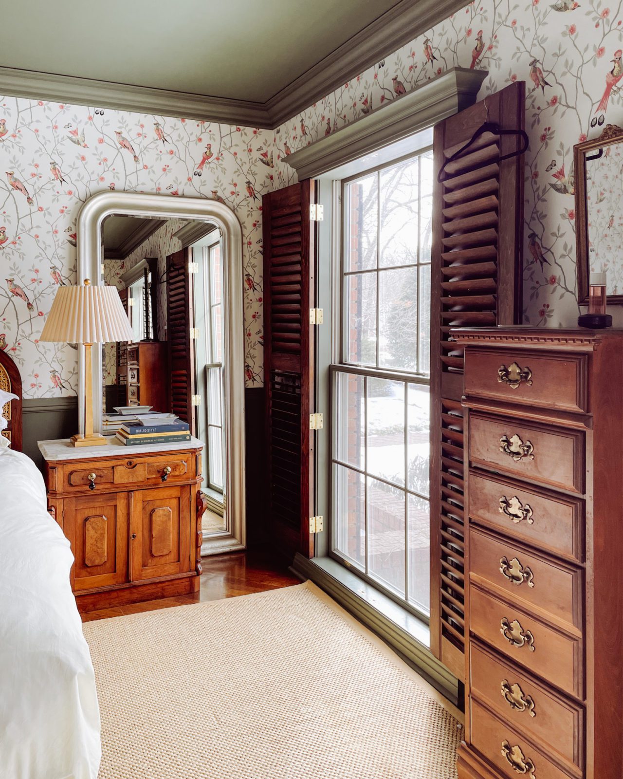 Main bedroom with bird wallpaper, green trim and ceiling, natural rug, wood shutters, IKEA floor mirror, and antique nightstands.