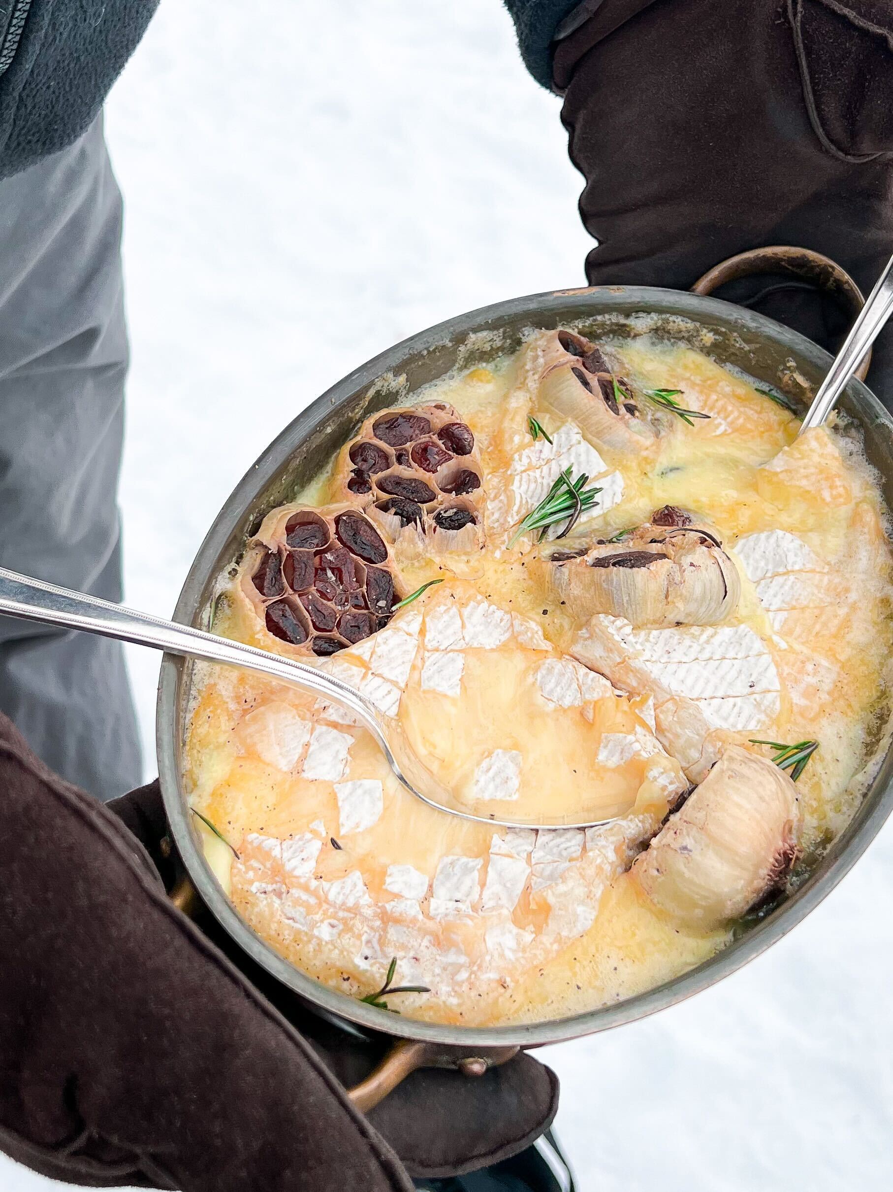 Two gloved hands are holding a rich baked dish of melted cheese, garlic, and rosemary