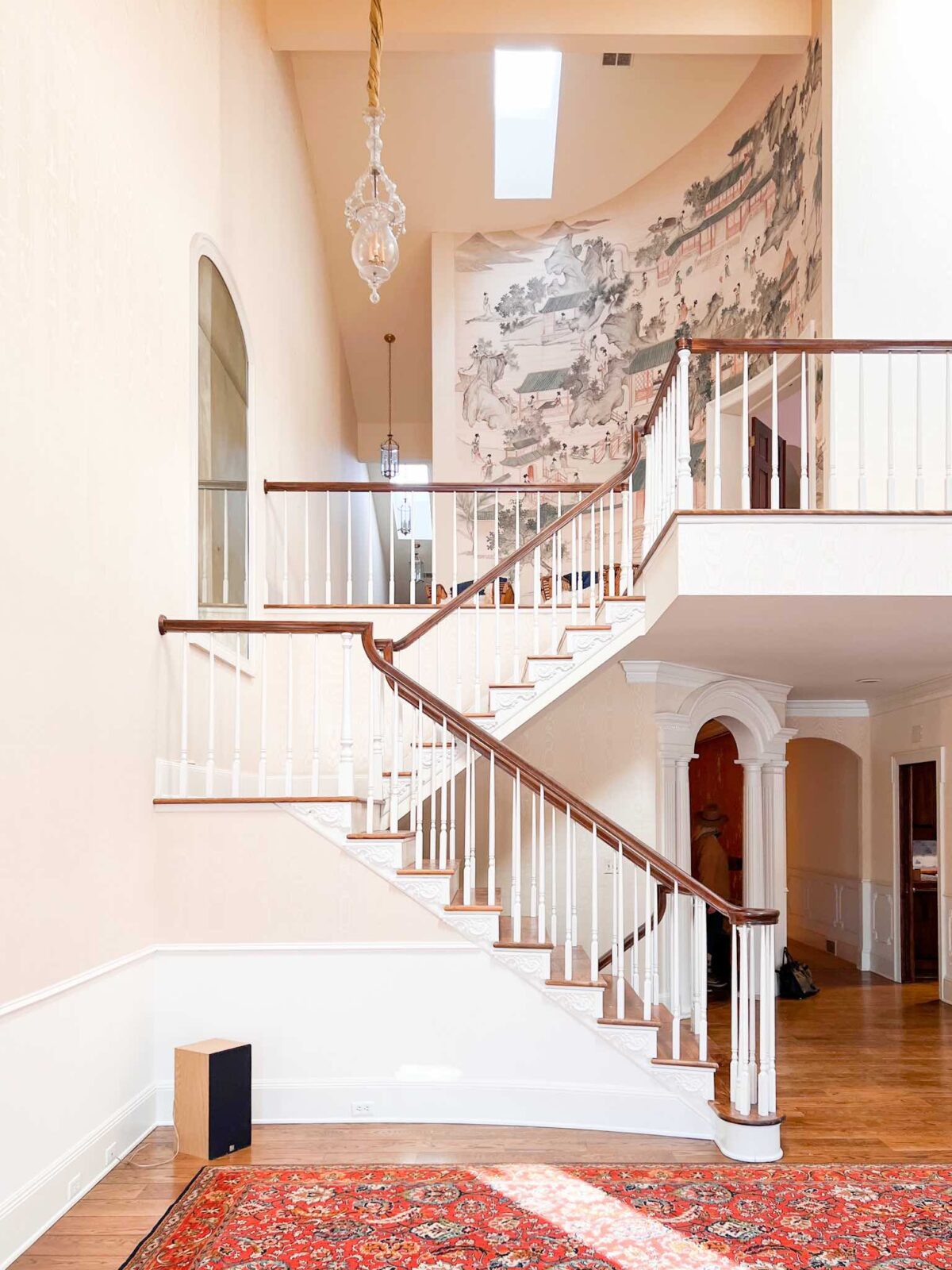 Two-story living room with a red rug on the lower level, a grand staircase, and a large mural on the upper level