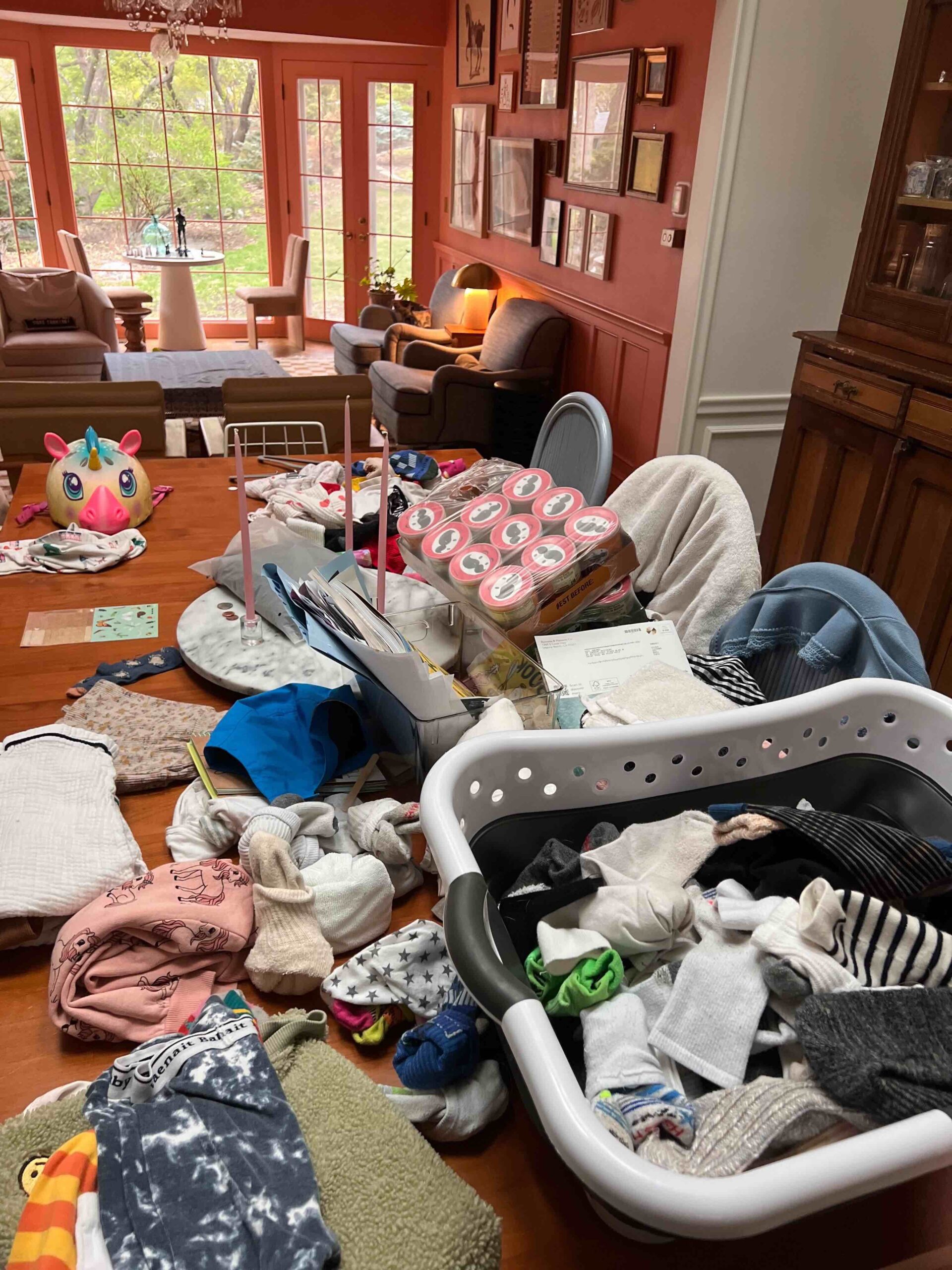 A dining room table is filled with the everyday messes of life: a laundry basket, scattered paper and mail, snacks, a bike helmet, and more