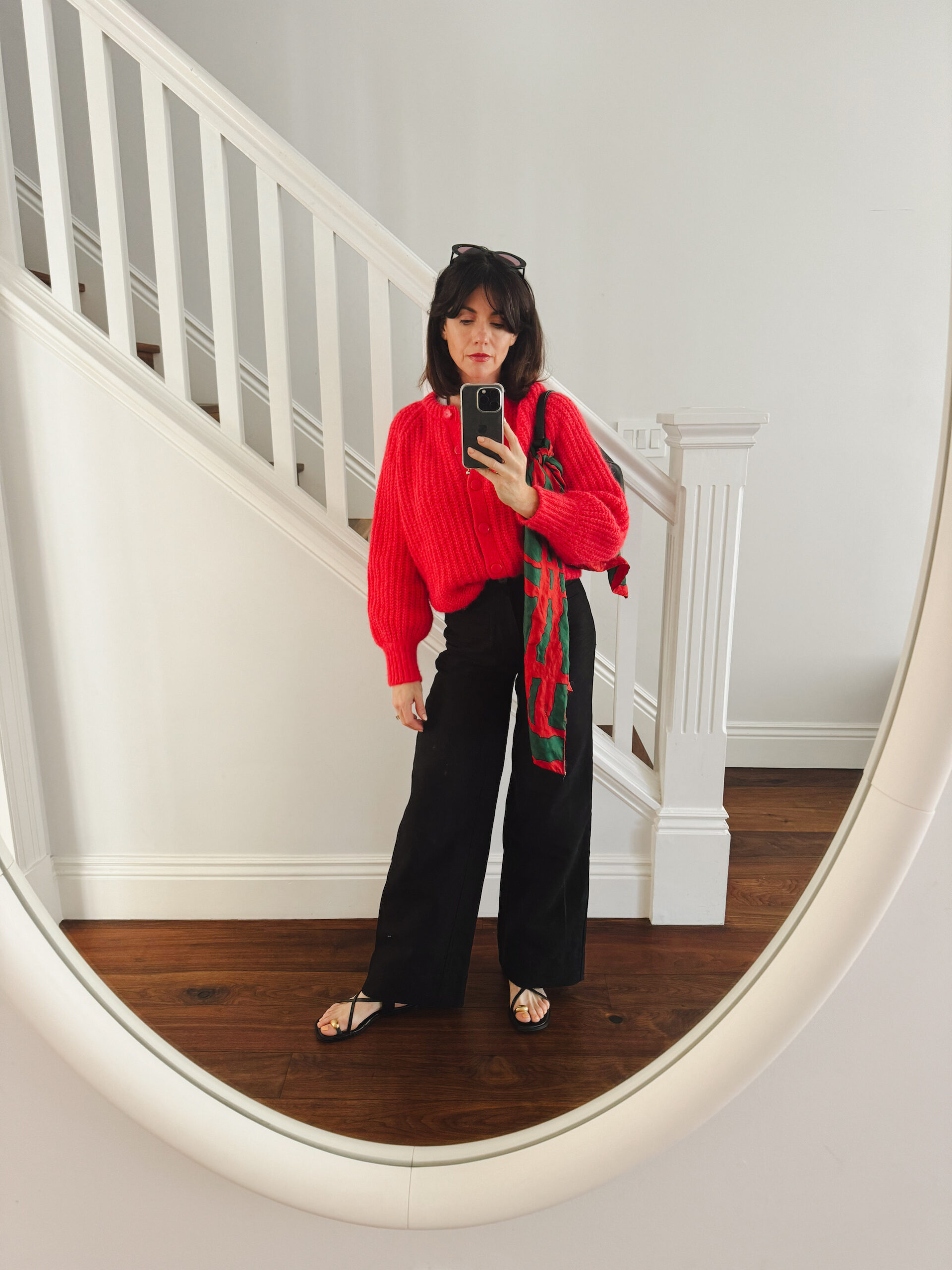 A woman stands in front of a staircase wearing a cherry red sweater, black trousers, black strappy sandals, and a black shoulder bag with a vintage red and green scarf tied on its handle.