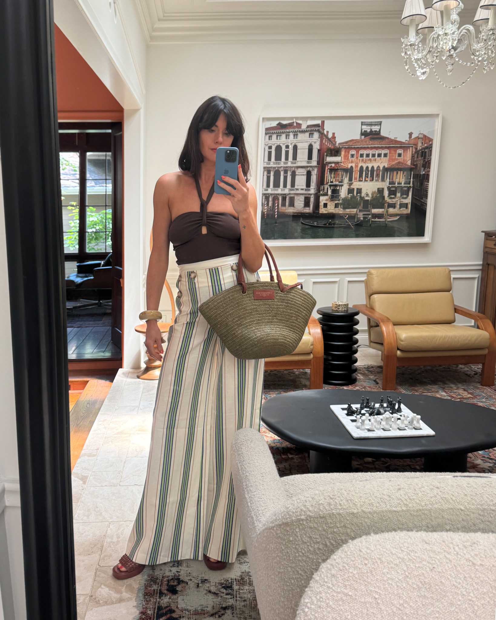 A woman wears a brown swimsuit with high-waisted, striped pants, brown heeled sandals, and a straw tote bag from Sézane