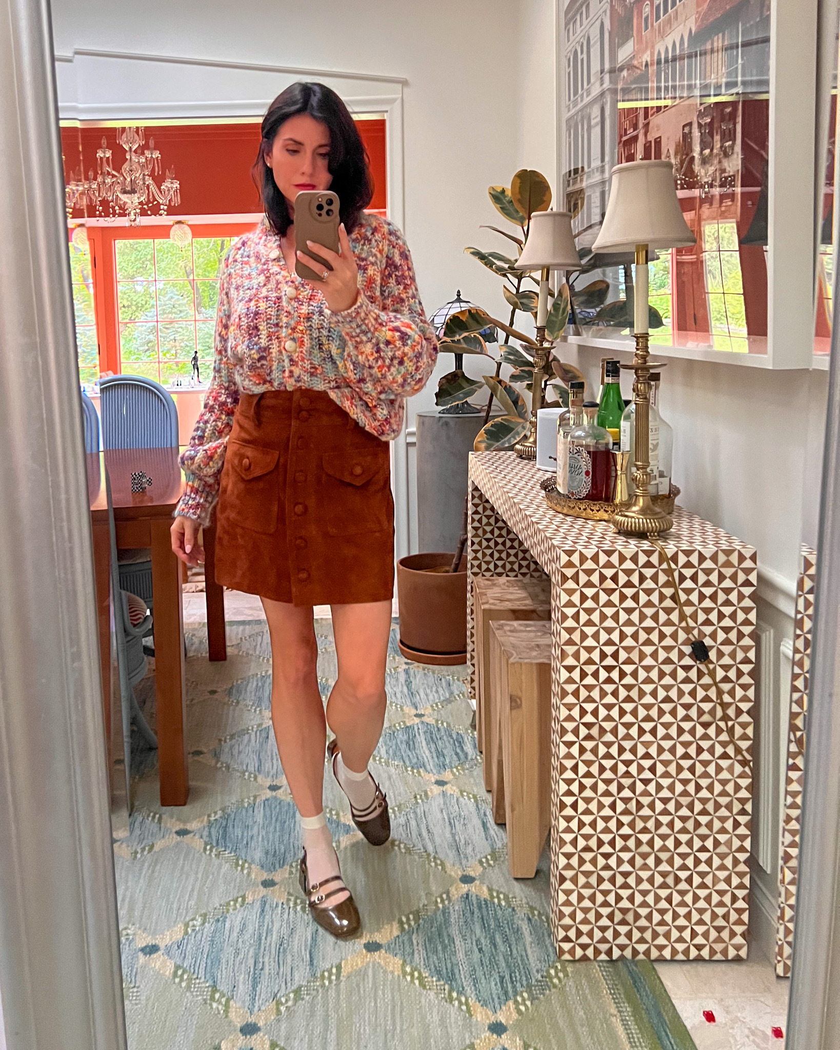 A mirror selfie of fall outfit consisting of a chunky colorful sweater, brown skirt, and olive flats