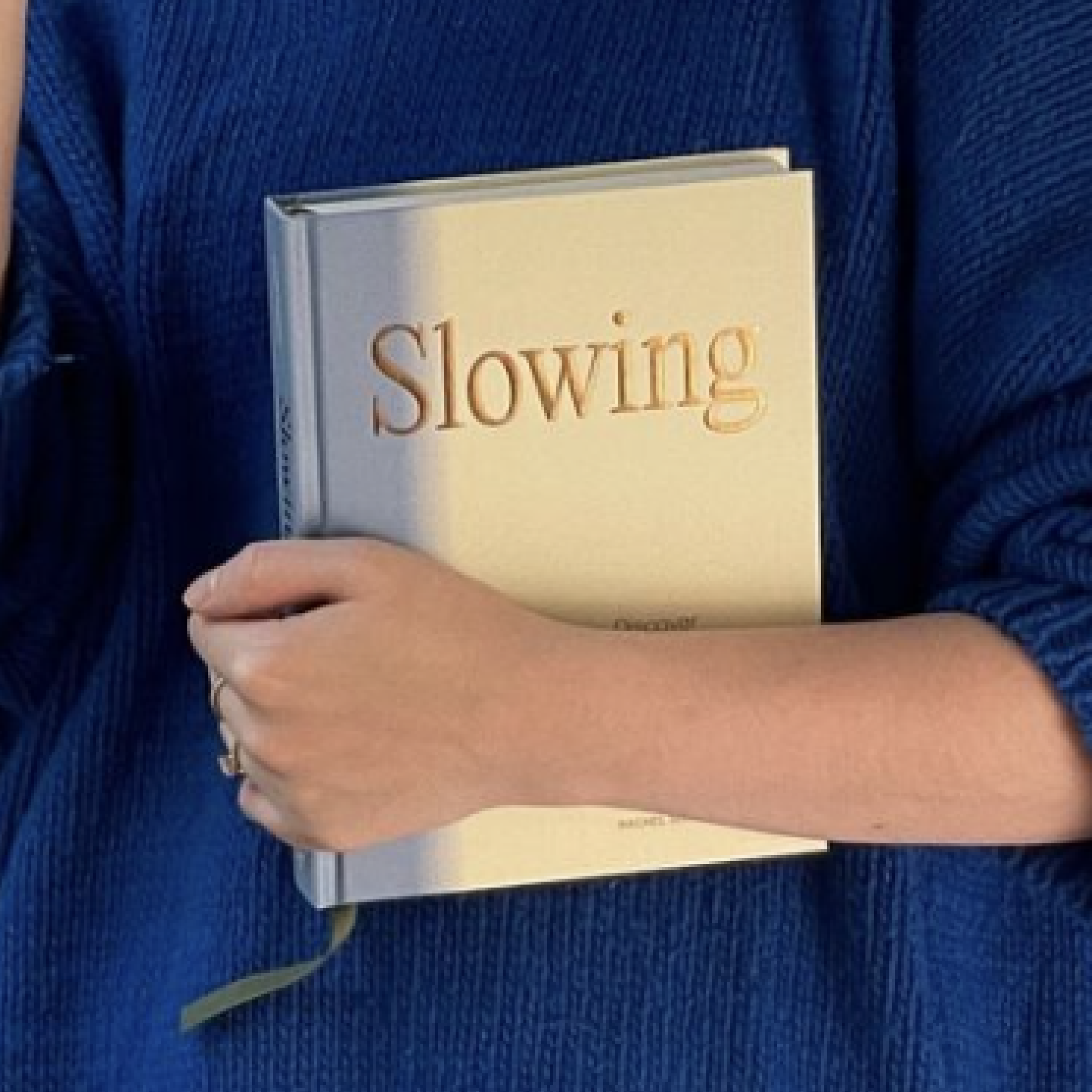 A woman is wearing a blue sweater and holding the book Slowing by Rachel Schwartzmann